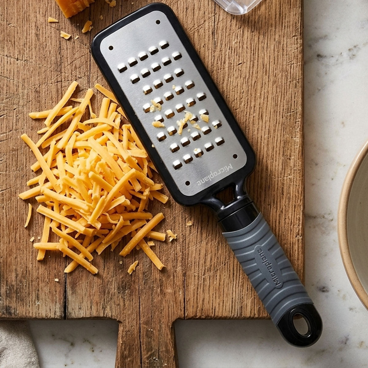 A stainless steel cheese grater with a black handle and gray grip, shown on a wooden cutting board next to shredded cheddar cheese on a marble surface.