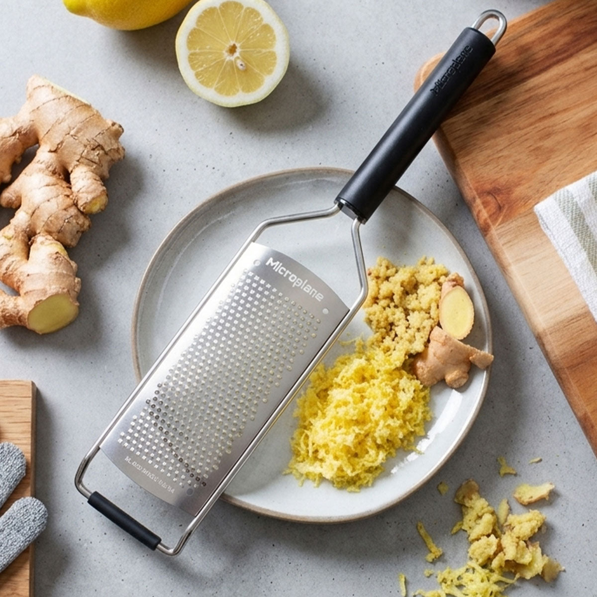 Microplane grater with grated lemon and ginger on a plate, surrounded by fresh lemons and ginger root.